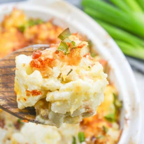 Close-up of a wooden spoon holding a portion of cheesy potato casserole, just like Grandma used to make, with a casserole dish and green onions in the background.