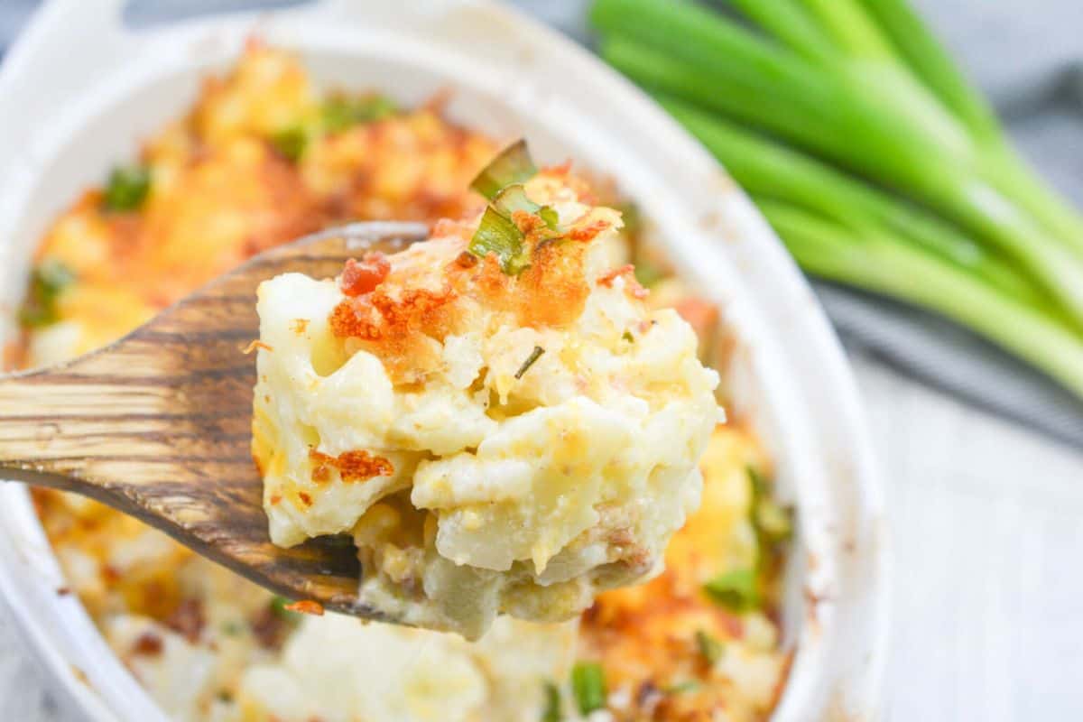 Close-up of a wooden spoon holding a portion of cheesy potato casserole, just like Grandma used to make, with a casserole dish and green onions in the background.
