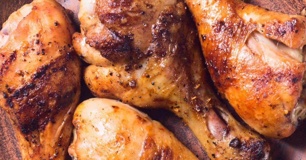 Four cooked, seasoned chicken drumsticks and thighs on a brown plate, showing crispy, golden-brown skin with visible grill marks—each piece bursting with flavor thanks to a savory marinade for grilled chicken drumsticks.