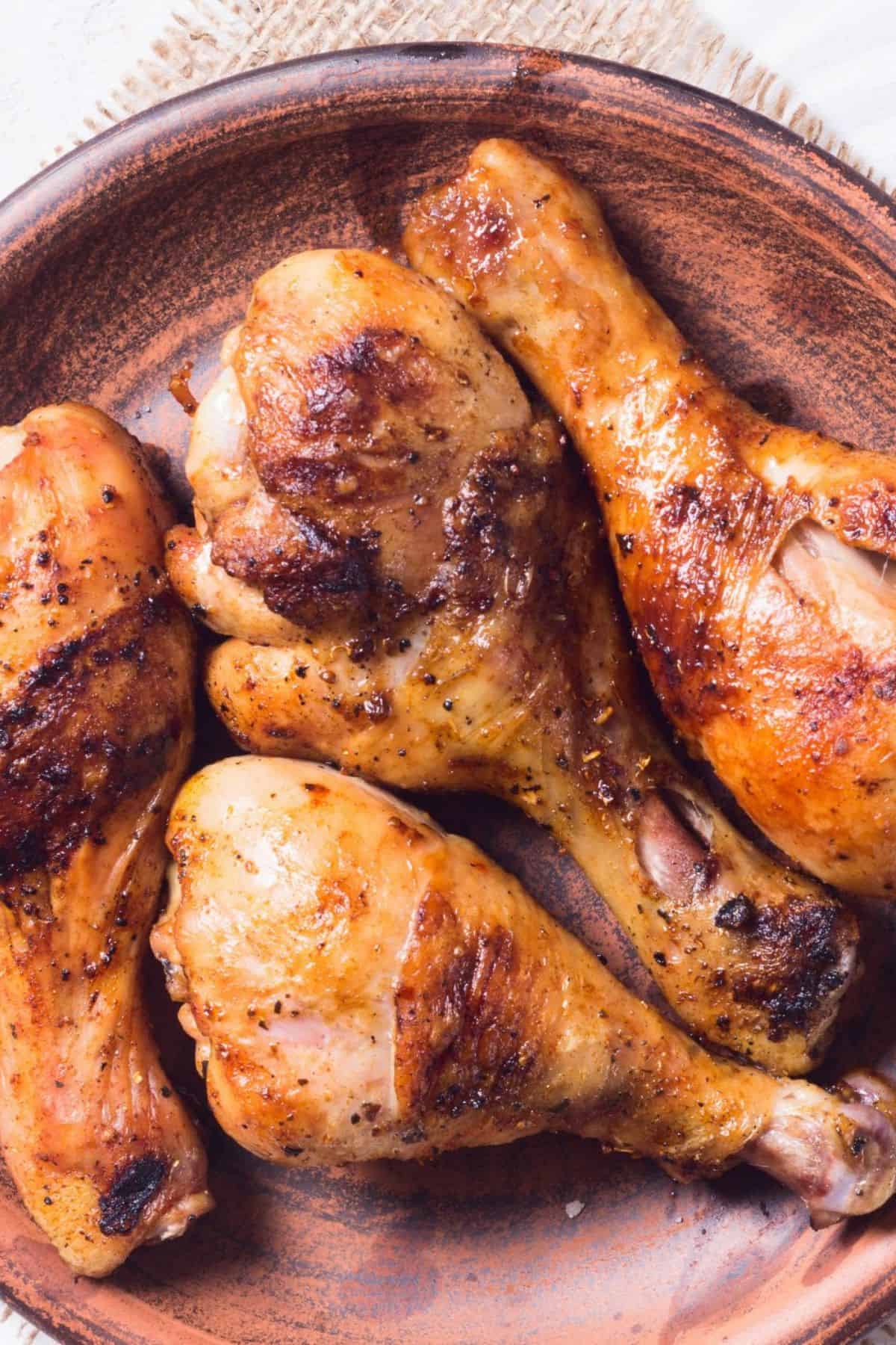 Four seasoned and roasted chicken drumsticks, infused with a flavorful marinade for grilled chicken drumsticks, are arranged on a brown ceramic plate.