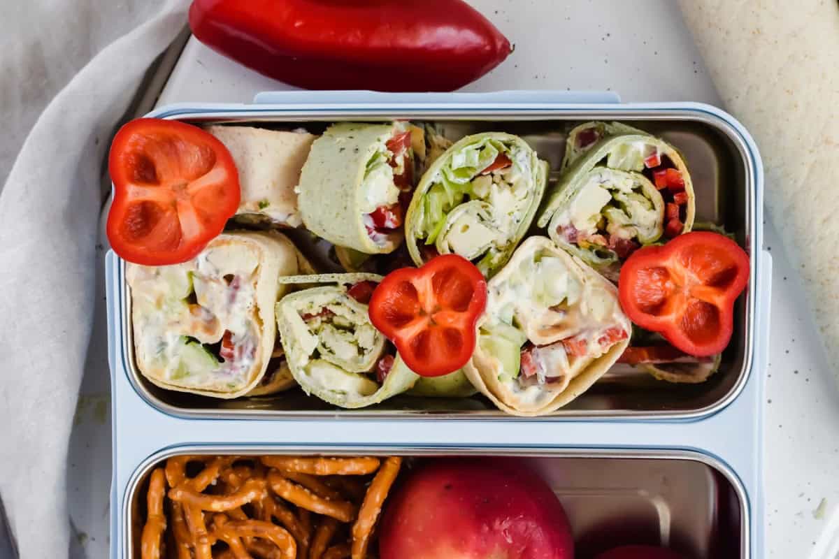 Sliced tortilla wraps, fresh lettuce, veggies, hummus and tzatziki sauce garnished with red bell pepper can be seen in the top compartment in a packable metal  and plastic lunchbox tray. Some pretzels and a peach can be seen in the bottom compartment while an uncut pepper rests outside and above the tray.