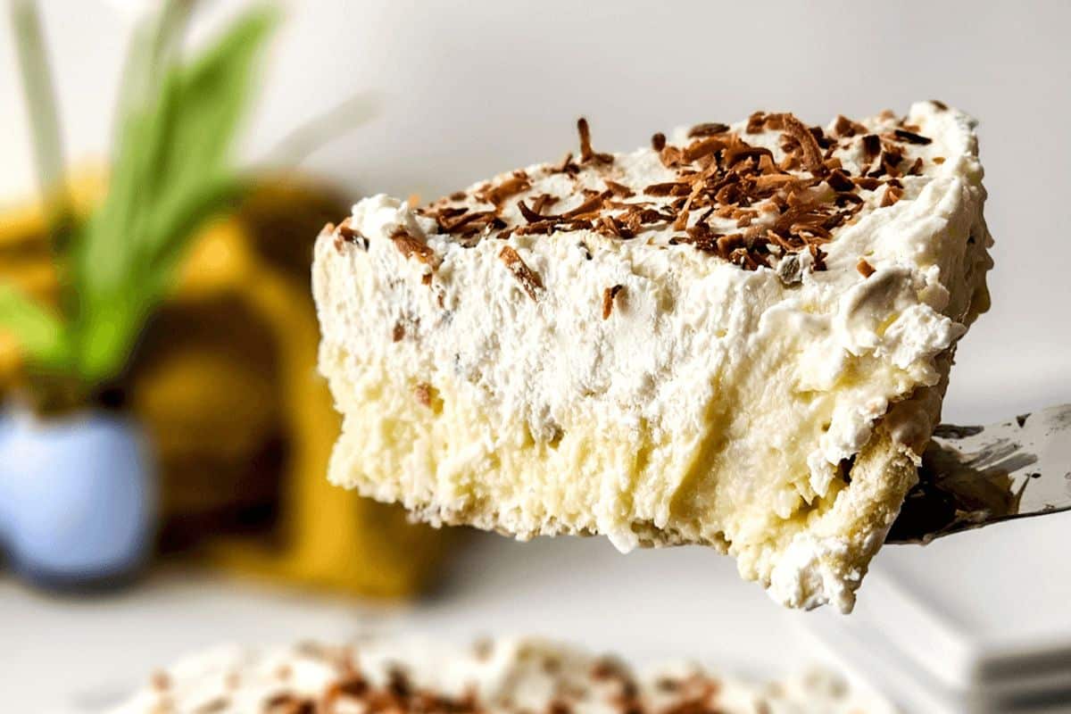 A slice of cream pie topped with chocolate shavings is being lifted from a plate. Reminiscent of old-fashioned dessert recipes, a plant and a blurred yellow object are in the background.