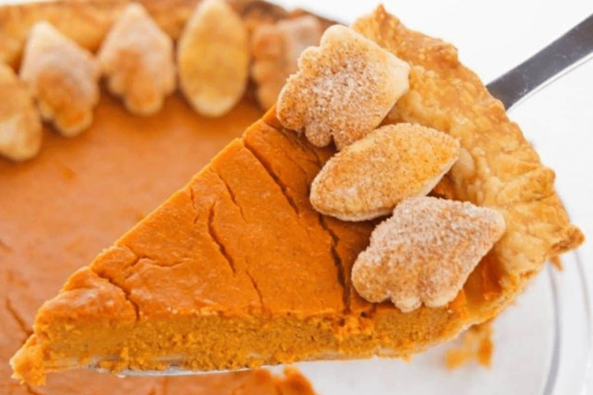 A slice of pumpkin pie garnished with small, leaf-shaped cookies is being lifted from the whole pie—a timeless treat straight from old-fashioned dessert recipes.