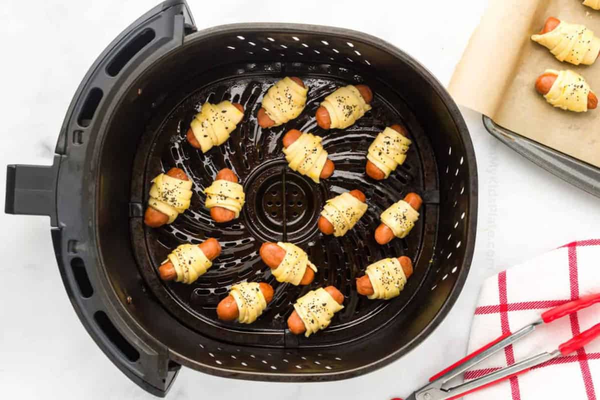 Mini hot dogs wrapped in crescent rolls and sprinkled with Everything But the Bagel seasoning rest in a black air fryer basket. A baking sheet lined with parchment paper can be seen in the top right corner and has two more pigs in blanket on it. Red metal tongs rest on a white and red checkered towel in the bottom right corner.
