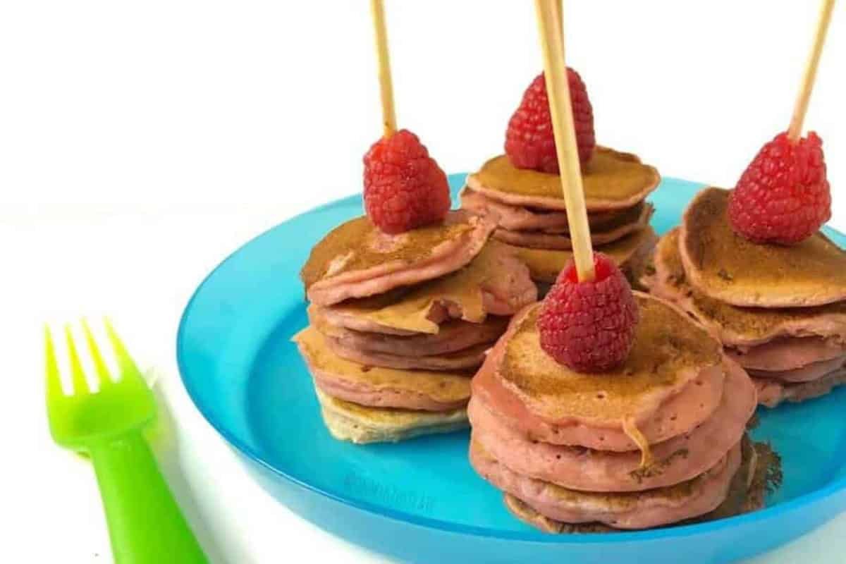 Four pink pancake kabobs topped with one raspberry each sit on a translucent blue plastic plate next to a translucent green plastic fork.