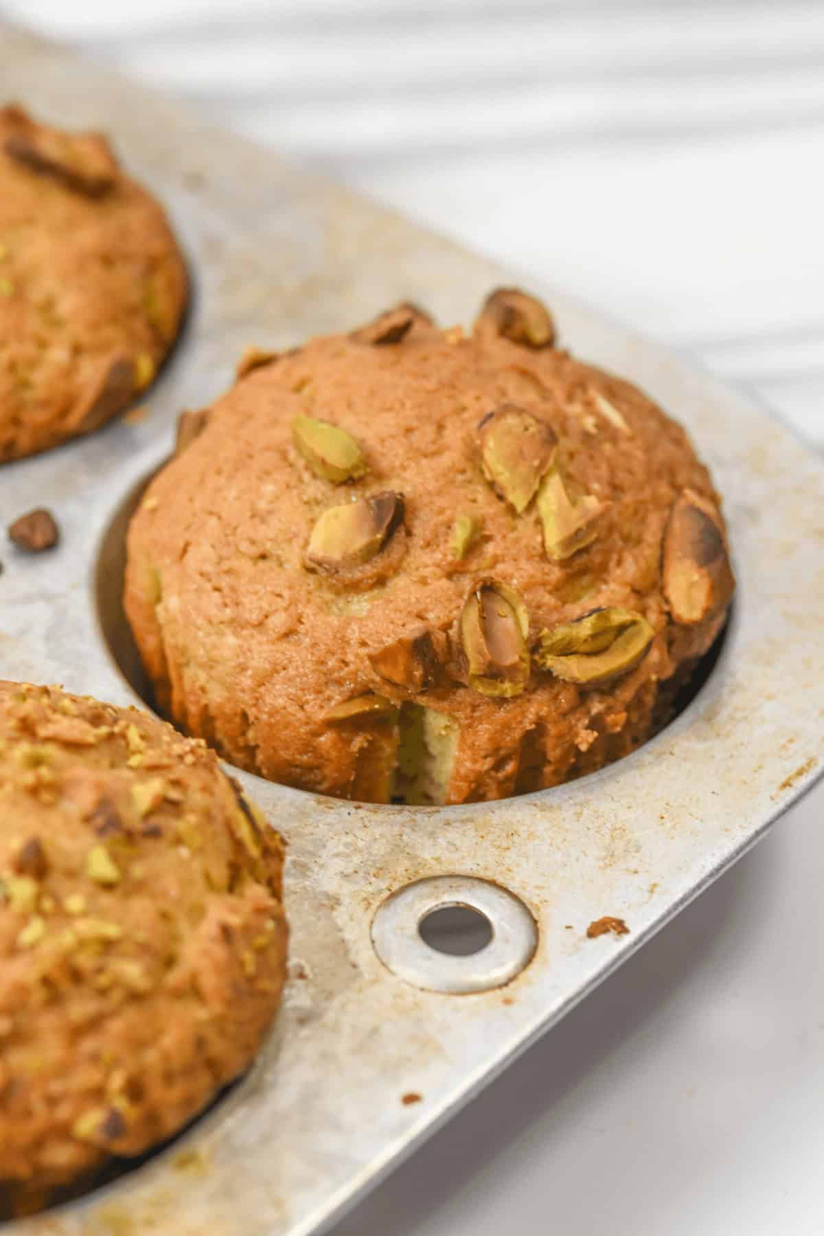 A close-up of freshly baked pistachio muffins nestles in a metal muffin tray, their golden tops enticing and aromatic, promising a delightful treat.