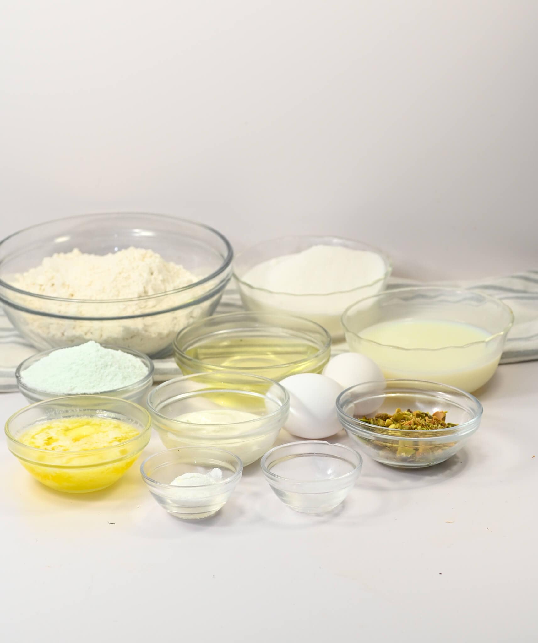 Various glass bowls filled with baking ingredients, including flour, sugar, eggs, melted butter, and milk—everything you need to whip up delightful muffins—are arranged on a white surface.