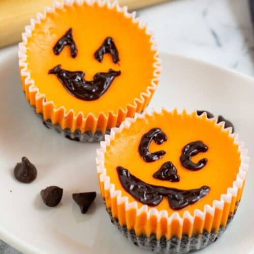 Two small orange cheesecakes with chocolate chip faces sit on a white plate, decorated with smiling jack-o-lantern designs. These charming Halloween desserts are perfect for adding a festive touch to your spooky celebrations.
