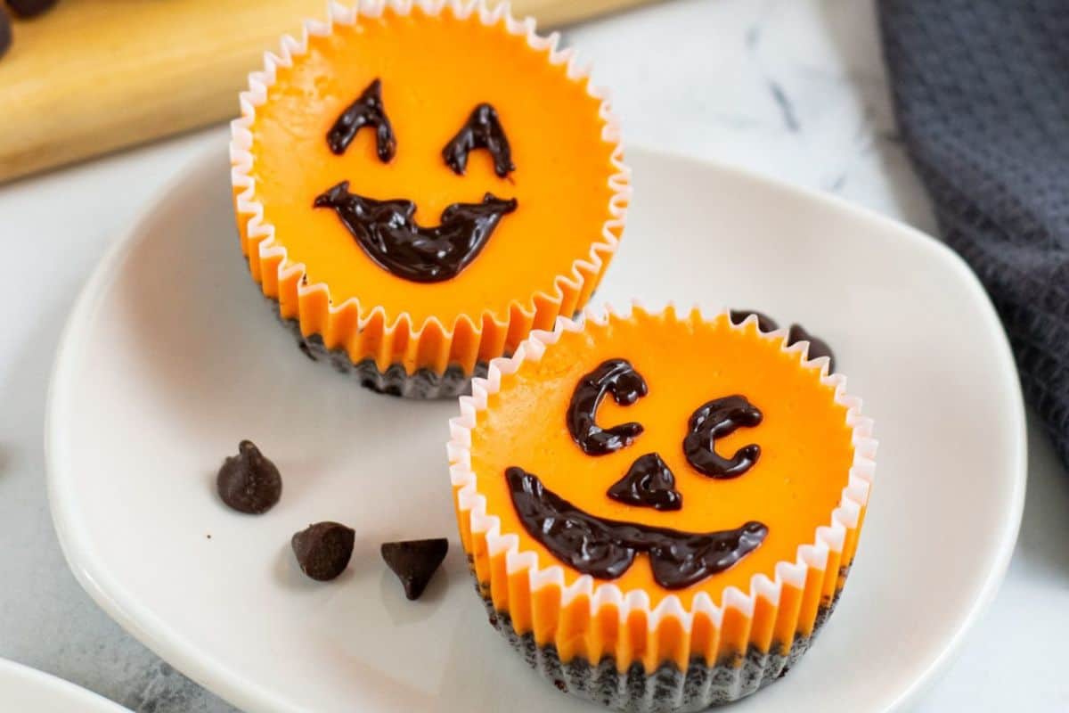 Two small orange cheesecakes with chocolate chip faces sit on a white plate, decorated with smiling jack-o-lantern designs. These charming Halloween desserts are perfect for adding a festive touch to your spooky celebrations.