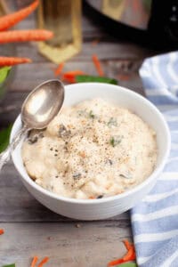 A bowl of creamy Slow Cooker Chicken Gnocchi Soup topped with herbs and black pepper, with a spoon resting on the edge. Nearby, a striped blue cloth and scattered carrot pieces lie on the wooden surface.