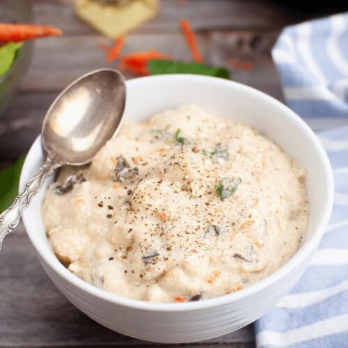 A bowl of creamy Slow Cooker Chicken Gnocchi Soup topped with herbs and black pepper, with a spoon resting on the edge. Nearby, a striped blue cloth and scattered carrot pieces lie on the wooden surface.