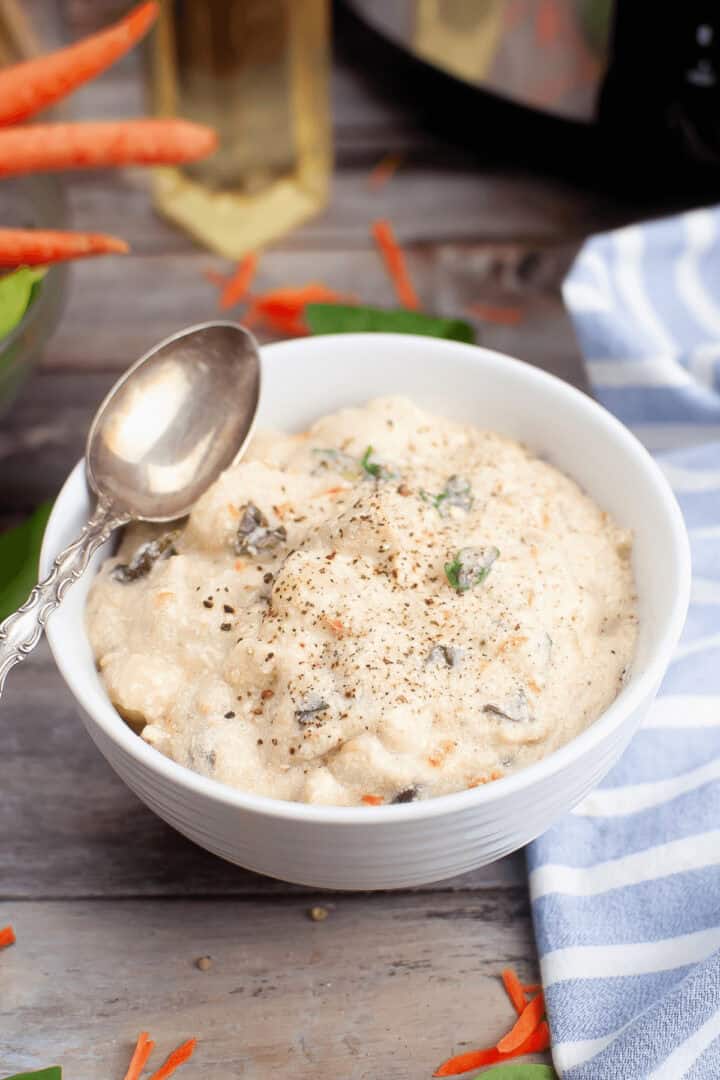 A bowl of creamy Slow Cooker Chicken Gnocchi Soup topped with herbs and black pepper, with a spoon resting on the edge. Nearby, a striped blue cloth and scattered carrot pieces lie on the wooden surface.