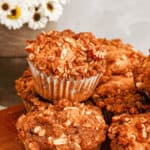 A stack of sweet potato muffins topped with chopped nuts sits on a wooden surface, with a bouquet of white daisies gently complementing the scene in the background.