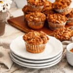 A stack of white plates with a single sweet potato muffin in front, other muffins on a wooden board behind. A small bowl of cinnamon is on the side.