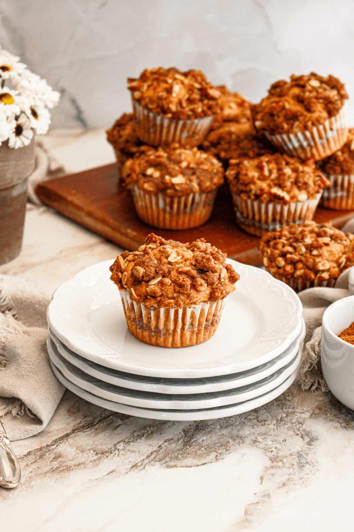 A stack of white plates with a single sweet potato muffin in front, other muffins on a wooden board behind. A small bowl of cinnamon is on the side.