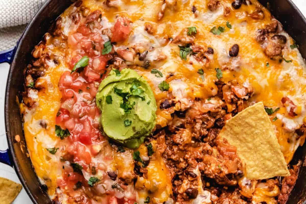 A close-up of a skillet filled with melted cheese, ground meat, black beans, tomatoes, guacamole, and a single tortilla chip evokes the comfort of 13 vintage recipes Grandma used to make.