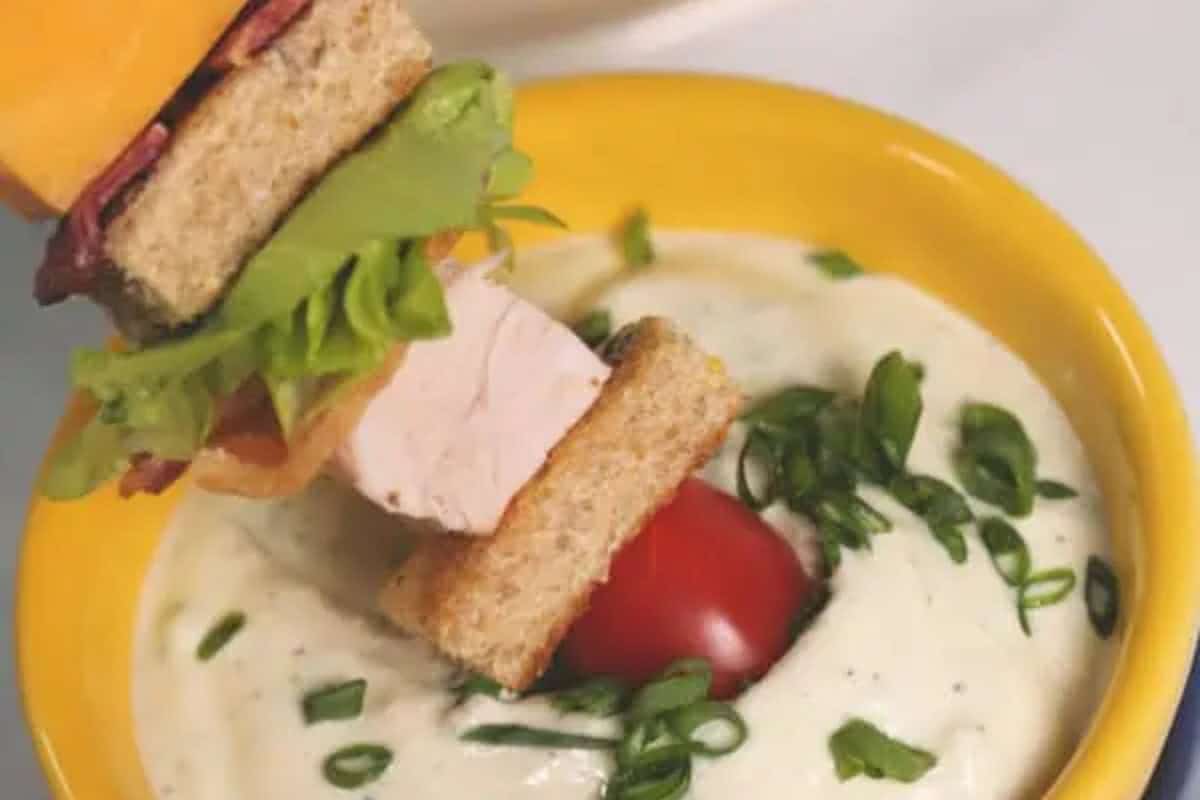 A skewer with turkey club sandwich pieces, lettuce, tomato, and cheese is dipped into a bowl of creamy dip topped with chopped green onions.