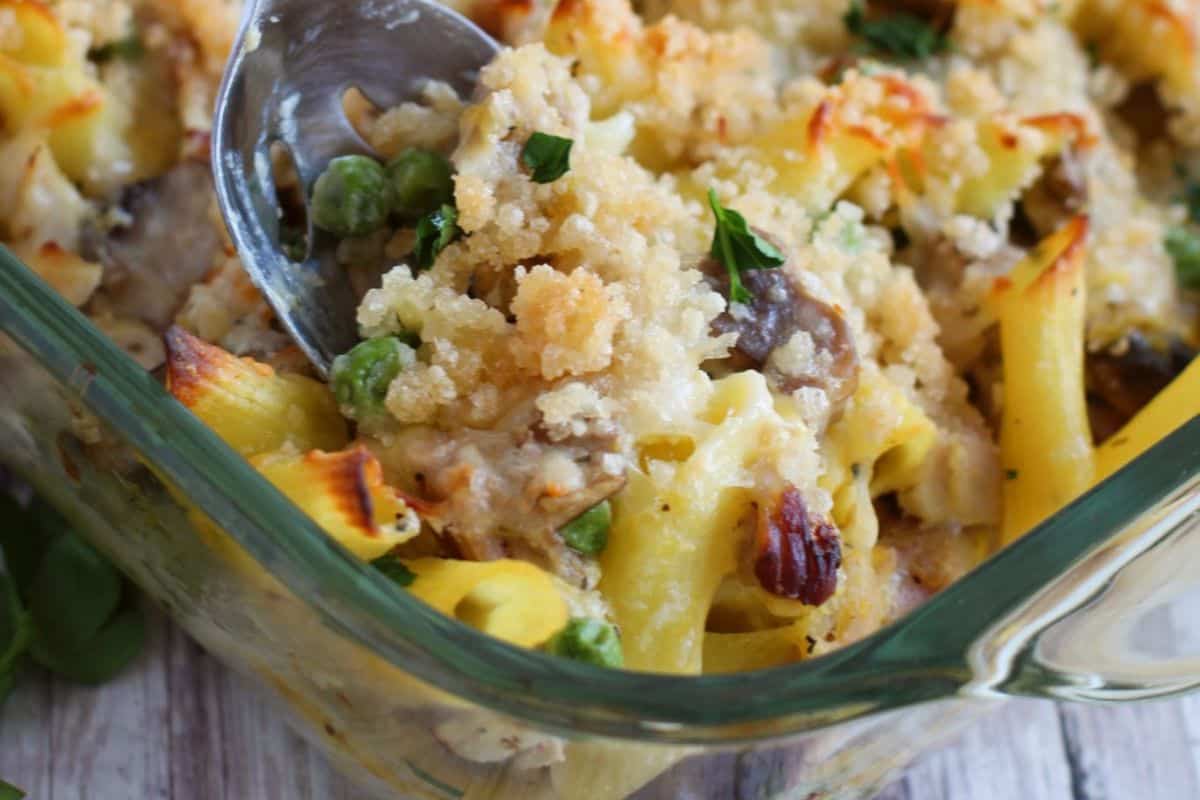 A close-up of a baked pasta casserole in a glass dish, featuring peas, mushrooms, and a golden breadcrumb topping reminiscent of vintage casserole recipes, being served with a metal spoon.