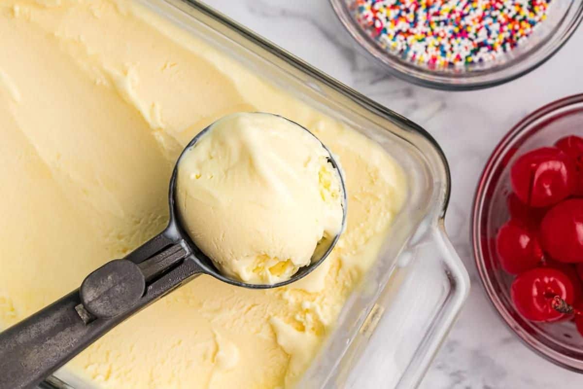 A metal ice cream scoop holds a scoop of vanilla ice cream over a glass container. Nearby, there are bowls of colorful sprinkles and red cherries, proving that old-fashioned dessert recipes never go out of style.
