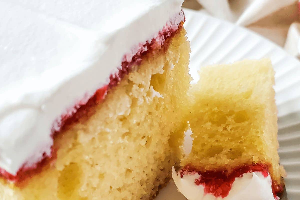 A slice of yellow cake with white frosting and a thin layer of red filling sits elegantly on a white plate, reminiscent of classic old-fashioned dessert recipes.