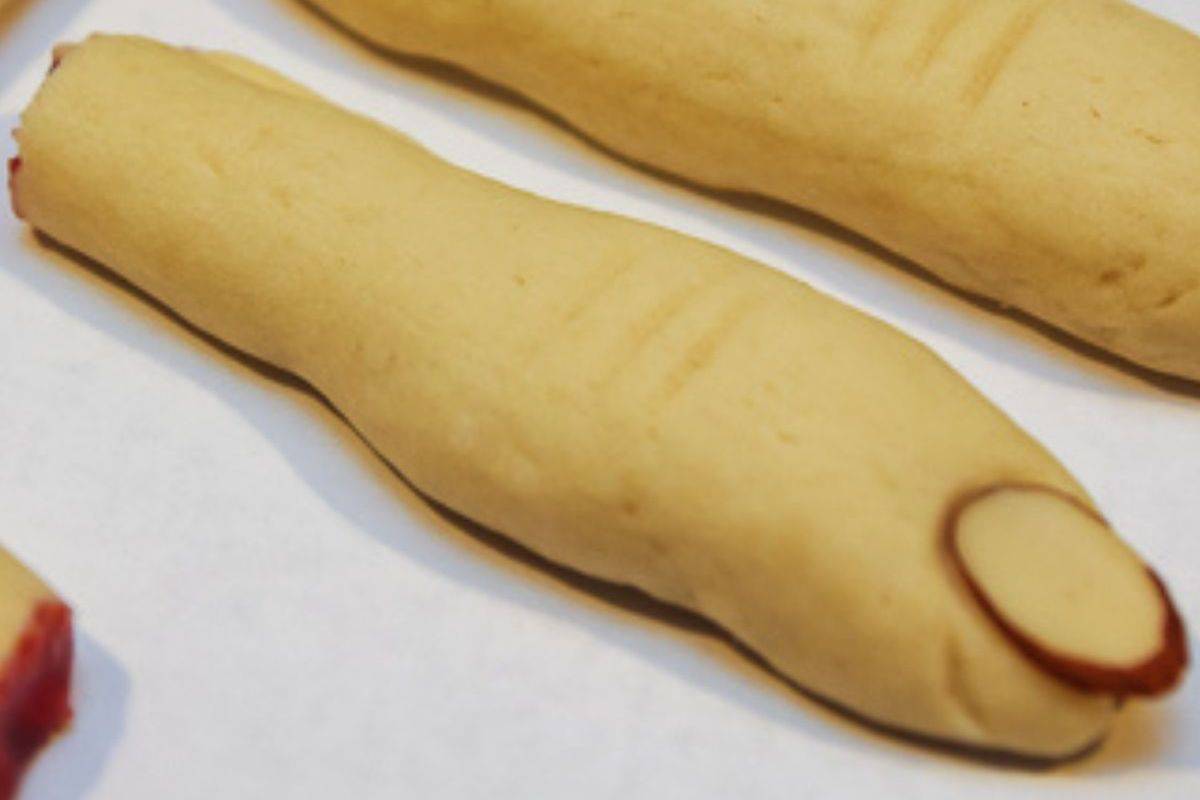 Close-up of a cookie shaped like a finger, with an almond slice representing a fingernail, placed on a white surface—perfect for spooky Halloween desserts.
