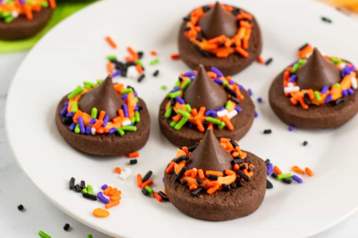 A white plate holds five chocolate cookies topped with chocolate kisses and sprinkled with colorful orange, green, purple, and black sprinkles. These Halloween treats are perfect for any spooky desserts spread, with more sprinkles scattered around the plate.