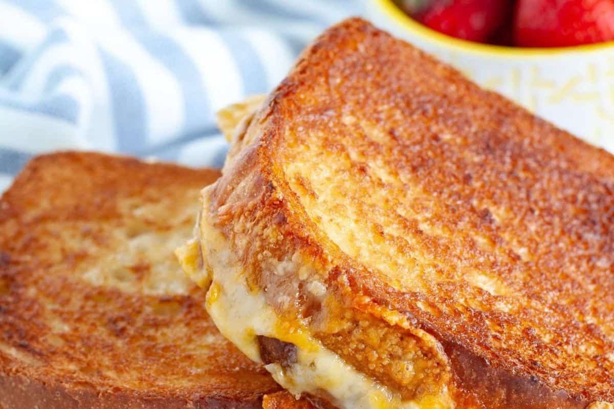 A close-up of two halves of a grilled cheese with a twist reveals melted cheese oozing from toasted bread. In the softly blurred background, a striped cloth and a bowl of strawberries add to the inviting scene.