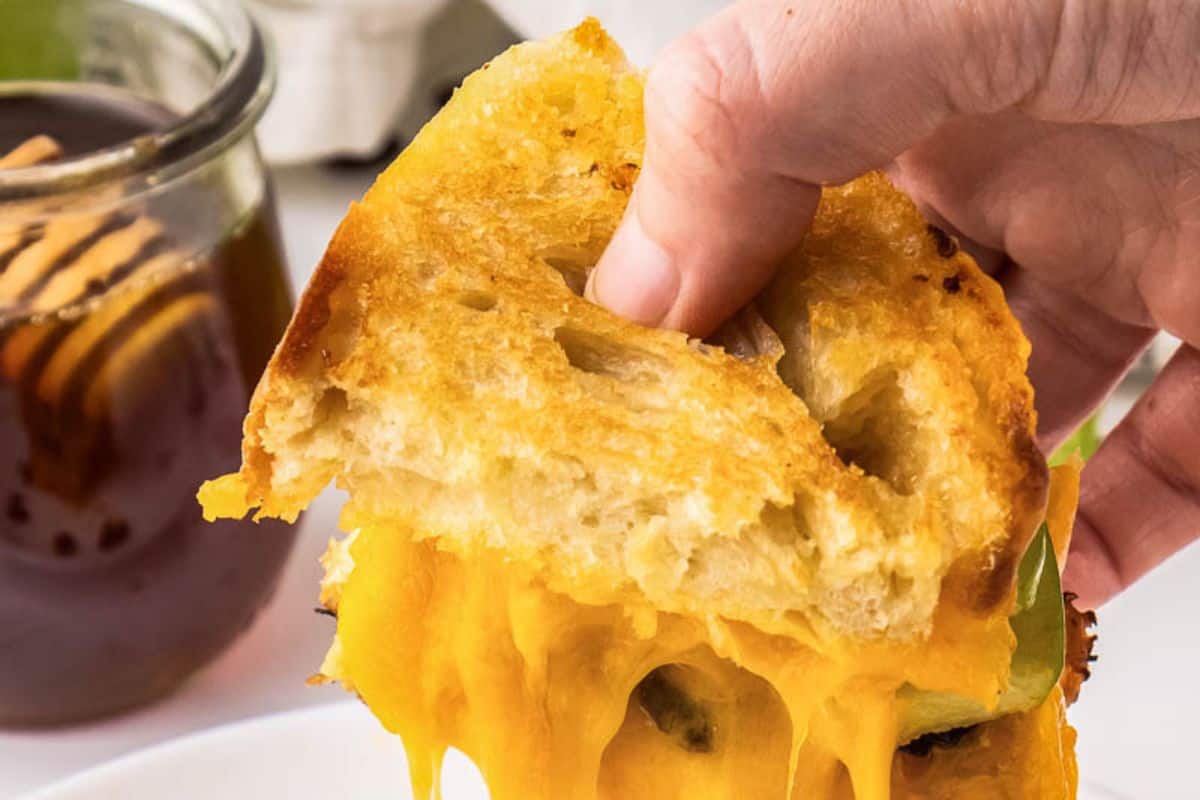 A hand holds a grilled cheese with a twist, its melted cheese stretching beside a jar of honey, where a wooden dipper rests invitingly.