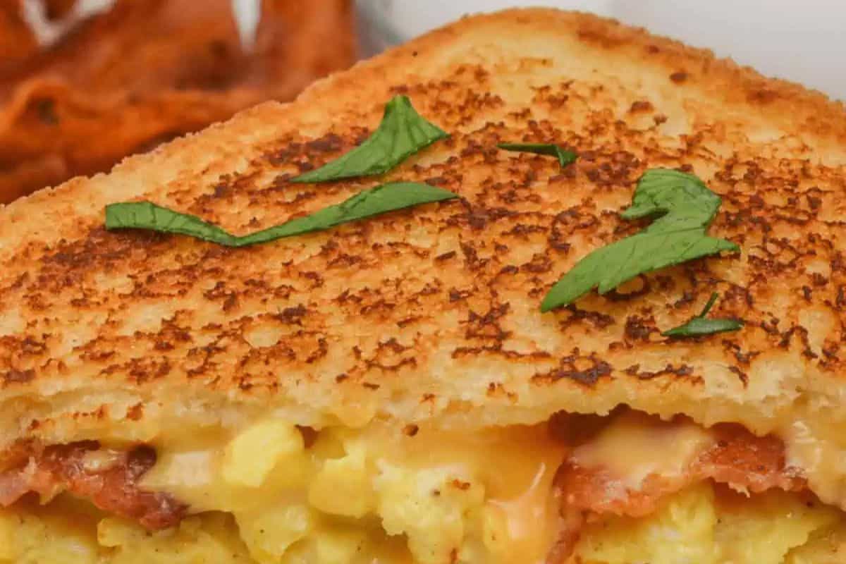 A delectable close-up of a grilled cheese with a twist, featuring scrumptious scrambled eggs, savory bacon, and a sprinkle of fresh green herbs on top.