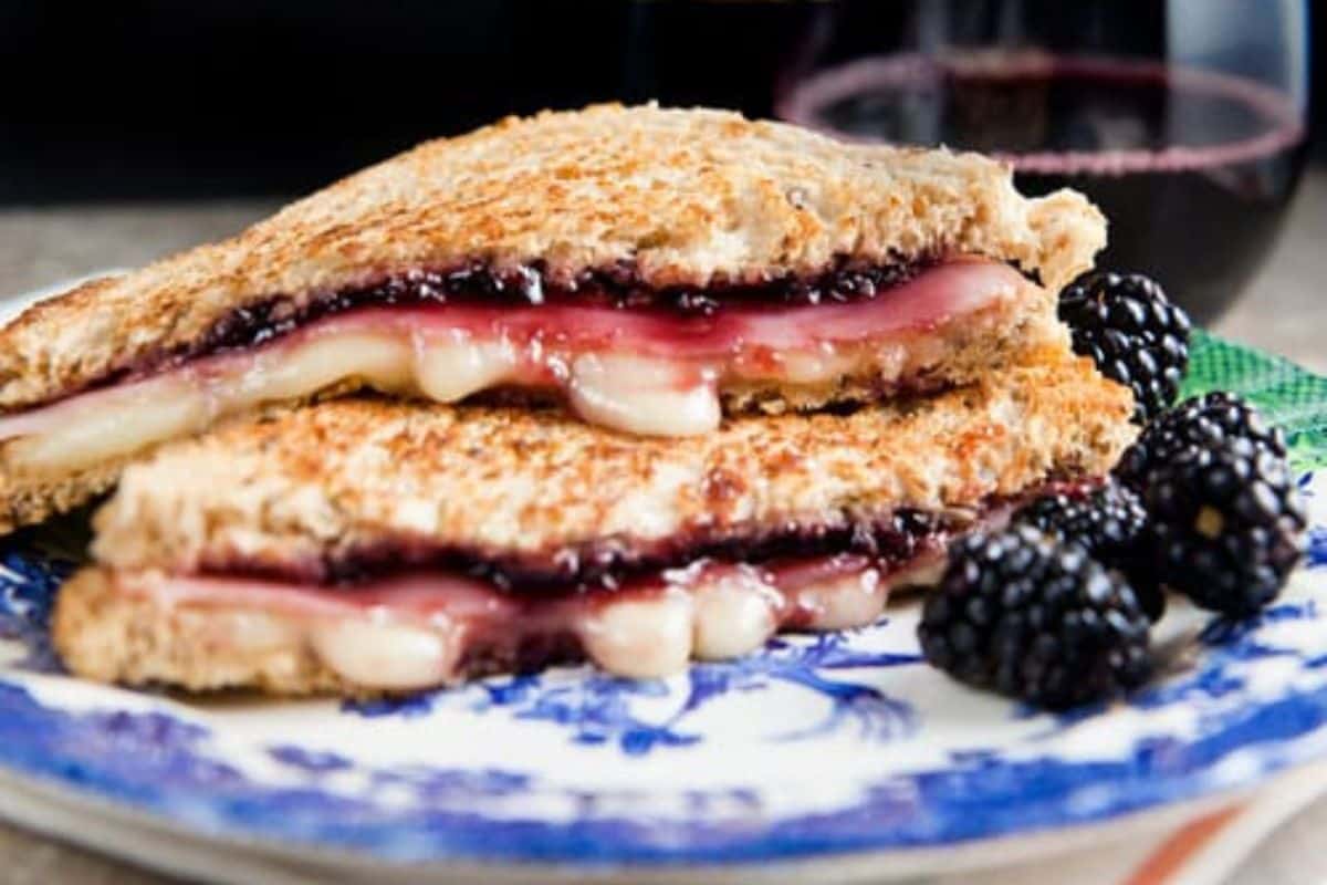 Grilled cheese with a twist: A crispy sandwich of melted cheese and berry jam, served on a plate with fresh blackberries. A glass of red wine is artfully placed in the background.