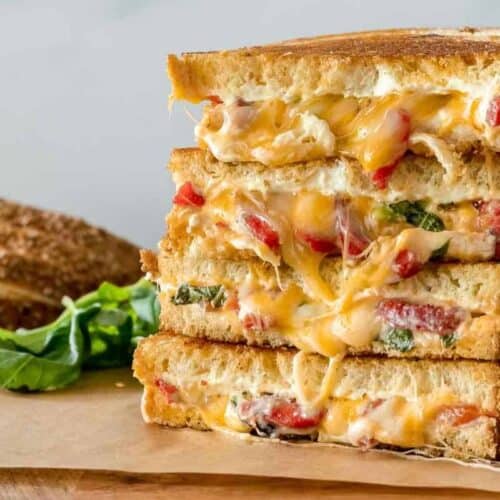 A stack of grilled cheese sandwiches with a twist, featuring melted cheese, tomatoes, and basil on a wooden cutting board, with slices of bread and fresh basil leaves in the background.