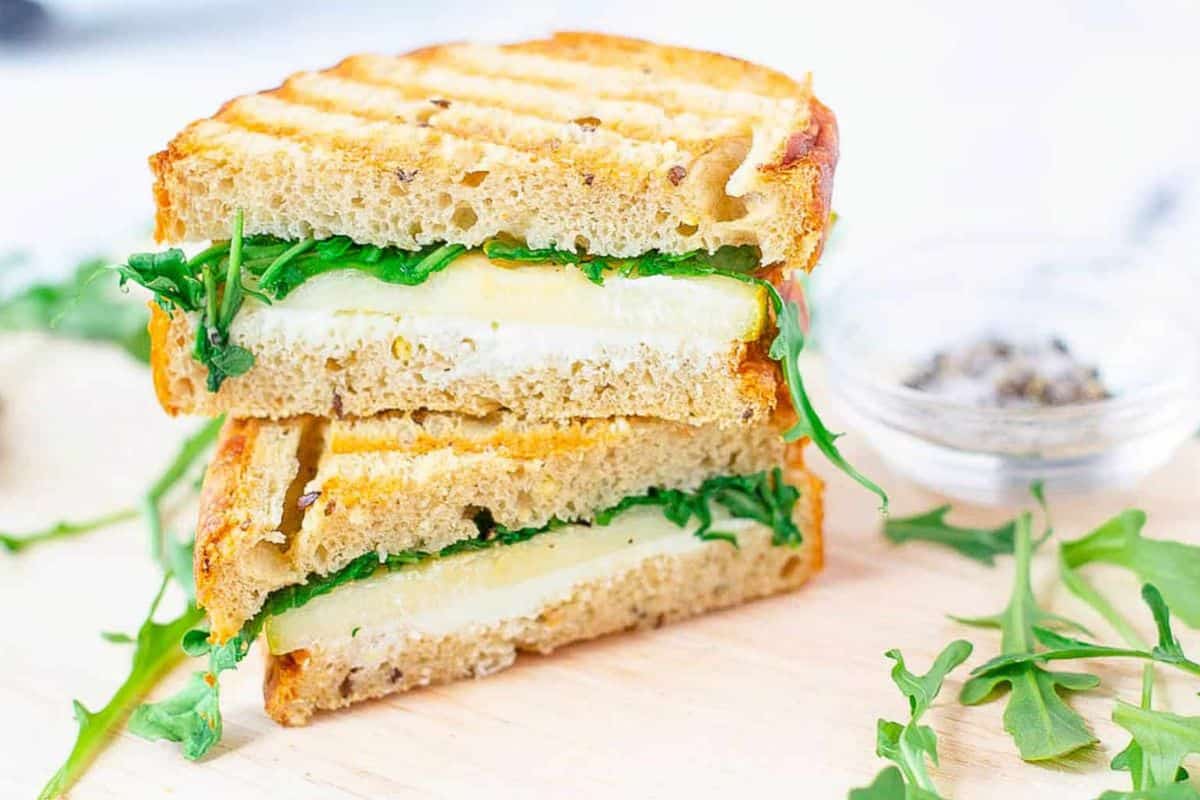 Two halves of a grilled cheese sandwich with a twist feature arugula and melted cheese on whole-grain bread, artfully stacked on a wooden surface. A dish with pepper sits in the background, adding an extra layer of flavor to this classic favorite.