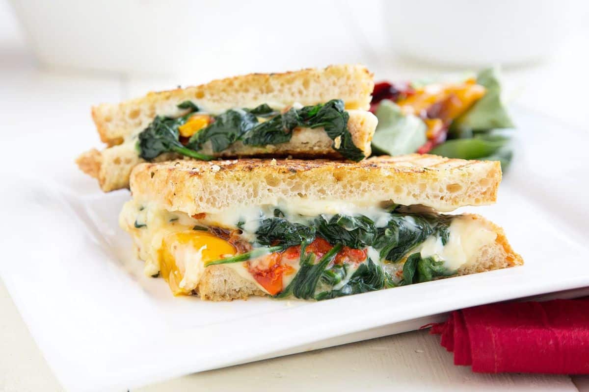 Two grilled cheese sandwiches with a twist—oozing melted cheese alongside fresh spinach and juicy tomatoes—sit invitingly on a white plate, garnished with mixed salad leaves.