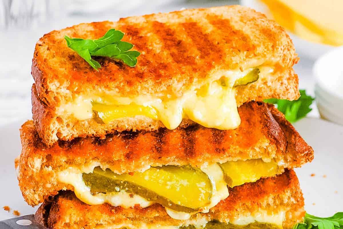 Grilled cheese with a twist: sandwiches stacked, filled with melted cheese and pickles, garnished with a parsley sprig.