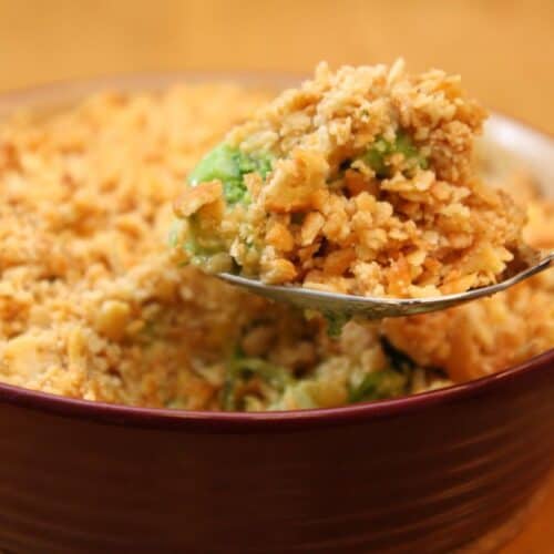 A close-up of a spoonful of casserole being lifted from a maroon dish. The casserole, part of the ultimate collection of broccoli recipes, has a crumbly, golden-brown topping and appears to contain green vegetables. Try it for your next family meal!
