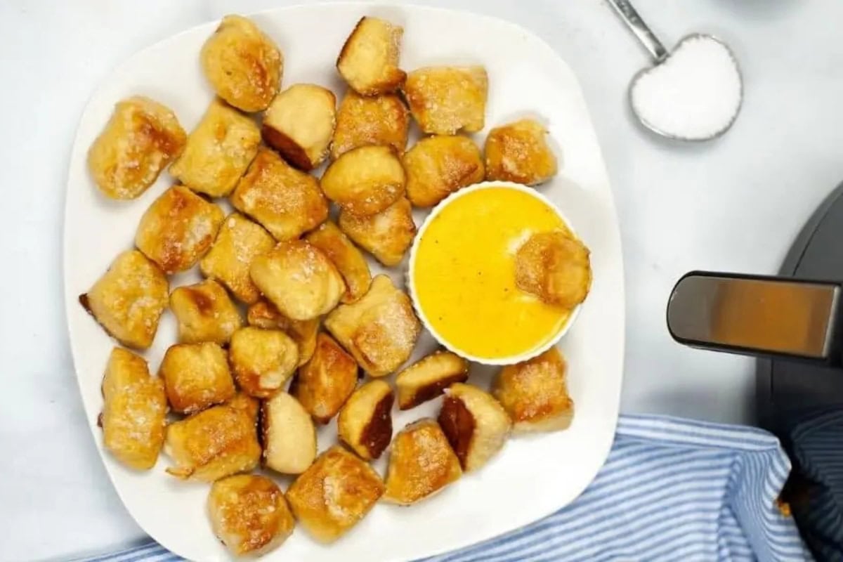 A plate of golden-brown pretzel bites with a small cup of yellow cheese dip, next to a spoonful of salt. The handle of an air fryer can be seen on the far right.