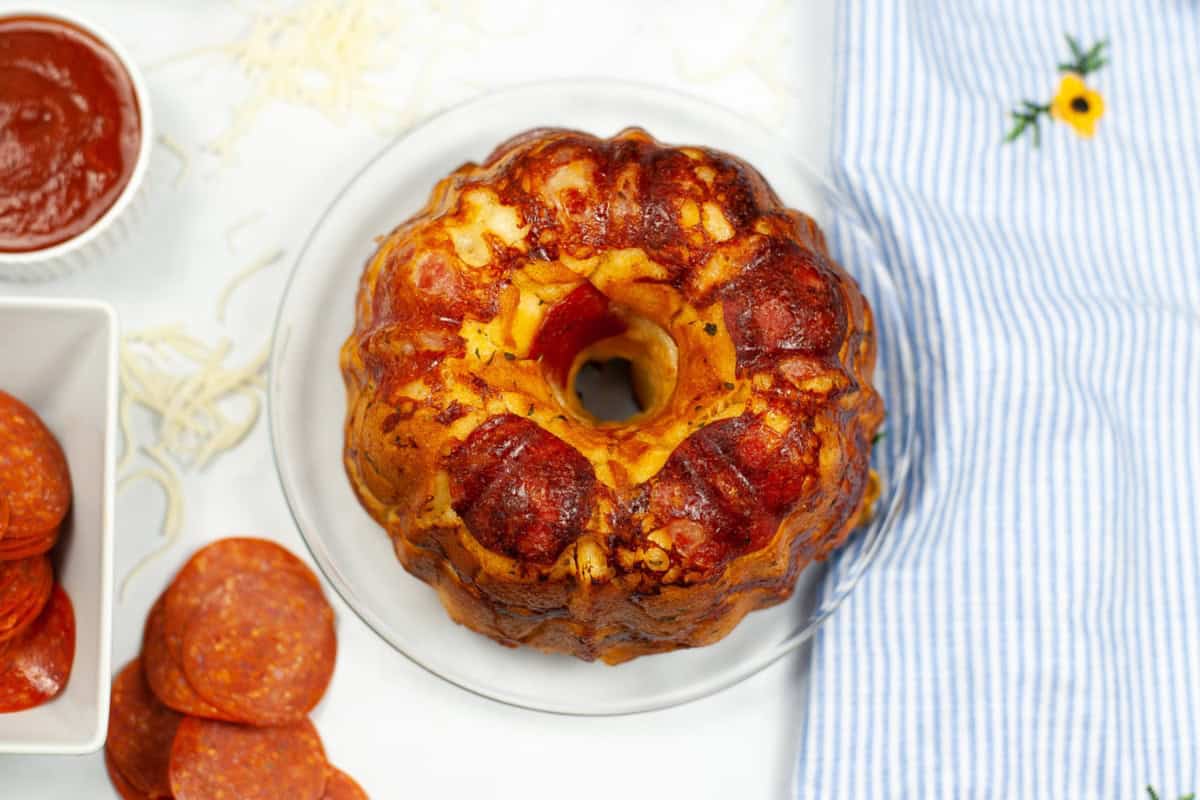 A baked pepperoni pull-apart bread ring on a glass plate, surrounded by slices of pepperoni, tomato sauce, shredded mozzarella, and a blue striped cloth.