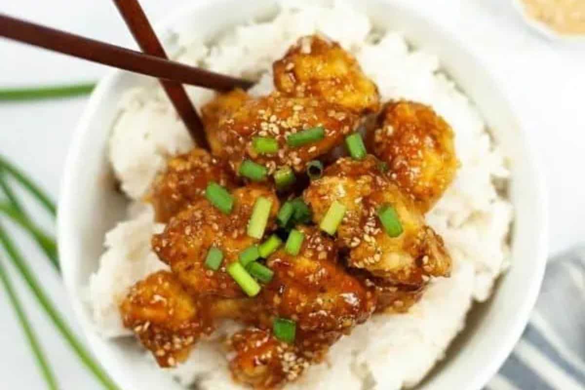 A bowl of white rice topped with glazed chicken pieces, garnished with chopped green onions and sesame seeds, with chopsticks on the side—a must try for fans of Asian dishes and better than takeout.