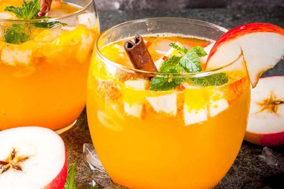 Two glasses of orange-colored apple mojito garnished with apple slices, mint leaves, and a cinnamon stick, and served over ice with fresh apple halves nearby.