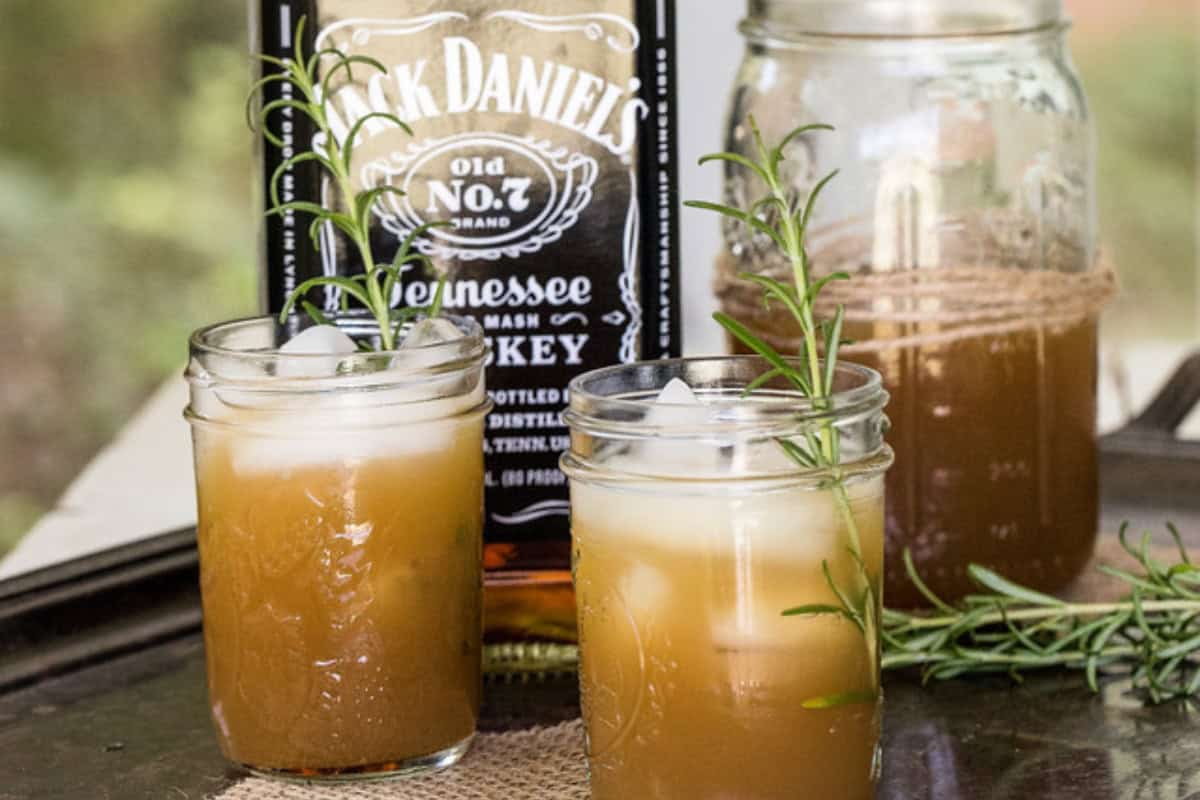 Two iced apple rosemary whiskey cocktails in mason jars garnished with rosemary, with a bottle of Jack Daniel’s whiskey and a jar of more cocktail in the background.