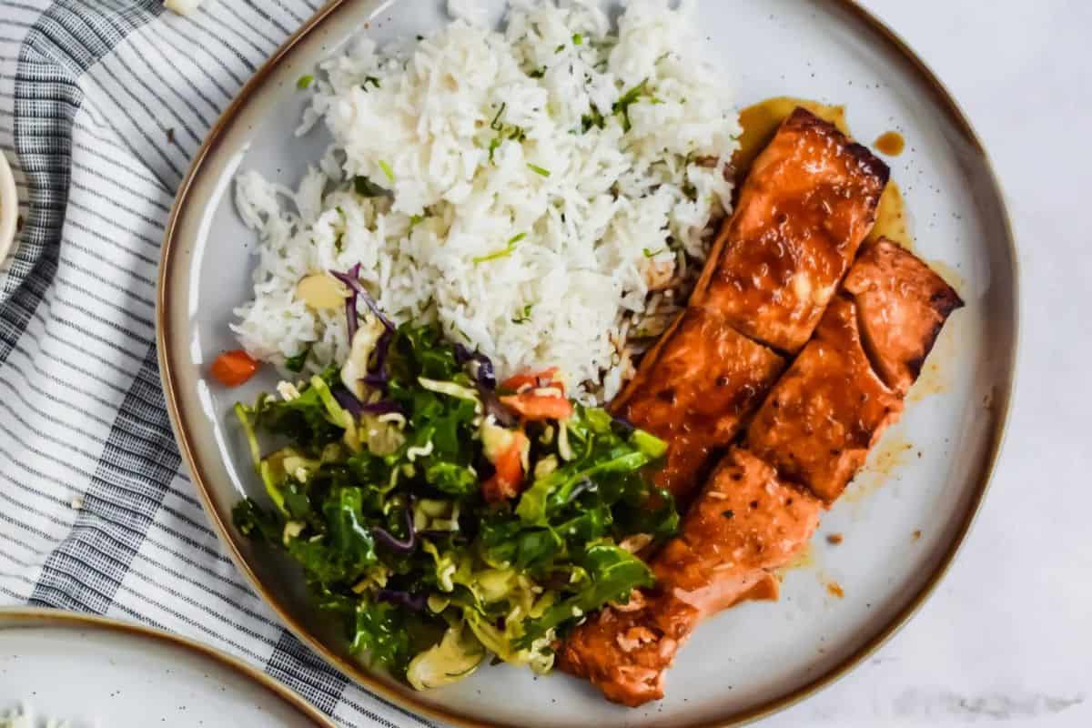 A plate with grilled salmon fillets, white rice, and a mixed green salad with chopped vegetables—this is a must try dish that's better than takeout and reminiscent of vibrant Asian dishes.