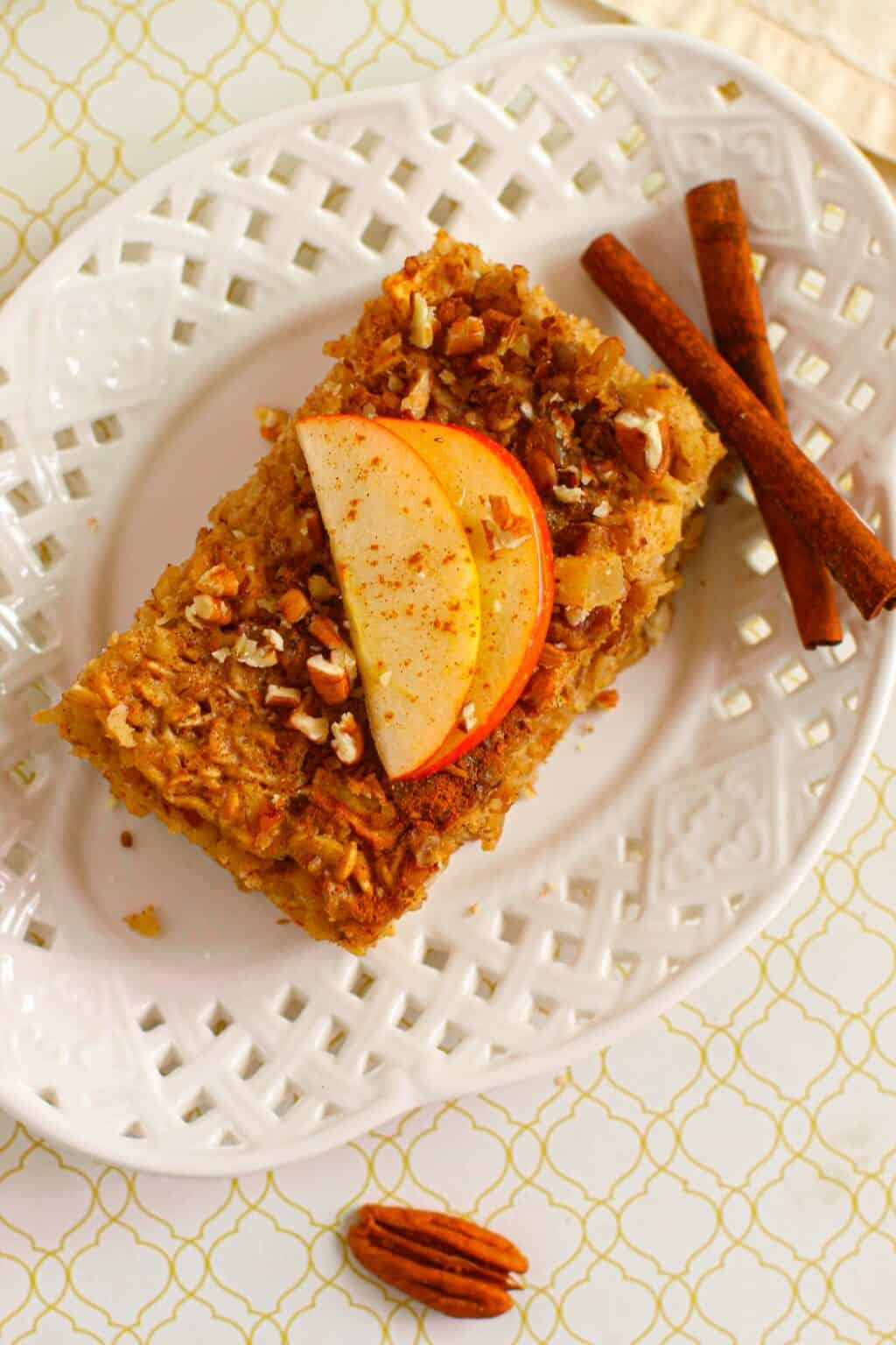 A slice of Baked Apple Oatmeal topped with apple slices and chopped nuts rests on a white plate, next to two cinnamon sticks and a pecan.
