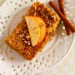A slice of Baked Apple Oatmeal topped with apple slices and chopped nuts rests on a white plate, next to two cinnamon sticks and a pecan.
