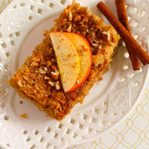 A slice of Baked Apple Oatmeal topped with apple slices and chopped nuts rests on a white plate, next to two cinnamon sticks and a pecan.