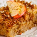 A Baked Apple Oatmeal square is topped with fresh apple slices, chopped nuts, and a sprinkle of cinnamon, served on a white plate with cinnamon sticks on the side.