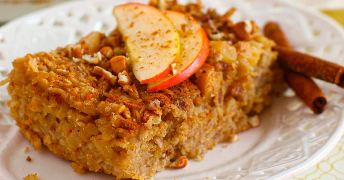 A Baked Apple Oatmeal square is topped with fresh apple slices, chopped nuts, and a sprinkle of cinnamon, served on a white plate with cinnamon sticks on the side.