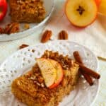 A slice topped with slices and pecans rests on a white plate, surrounded by cinnamon sticks.