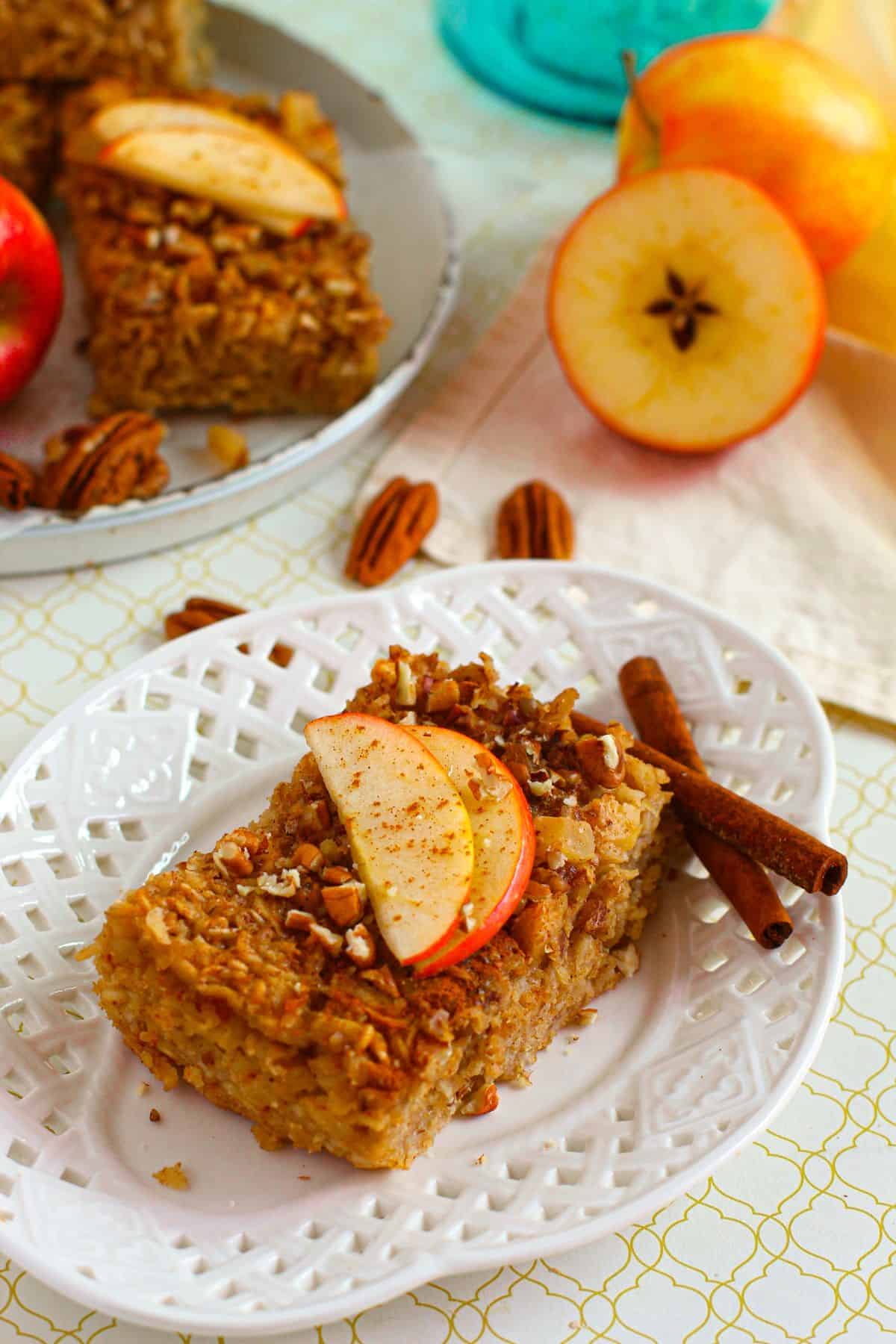 A slice  topped with slices and pecans rests on a white plate, surrounded by cinnamon sticks.