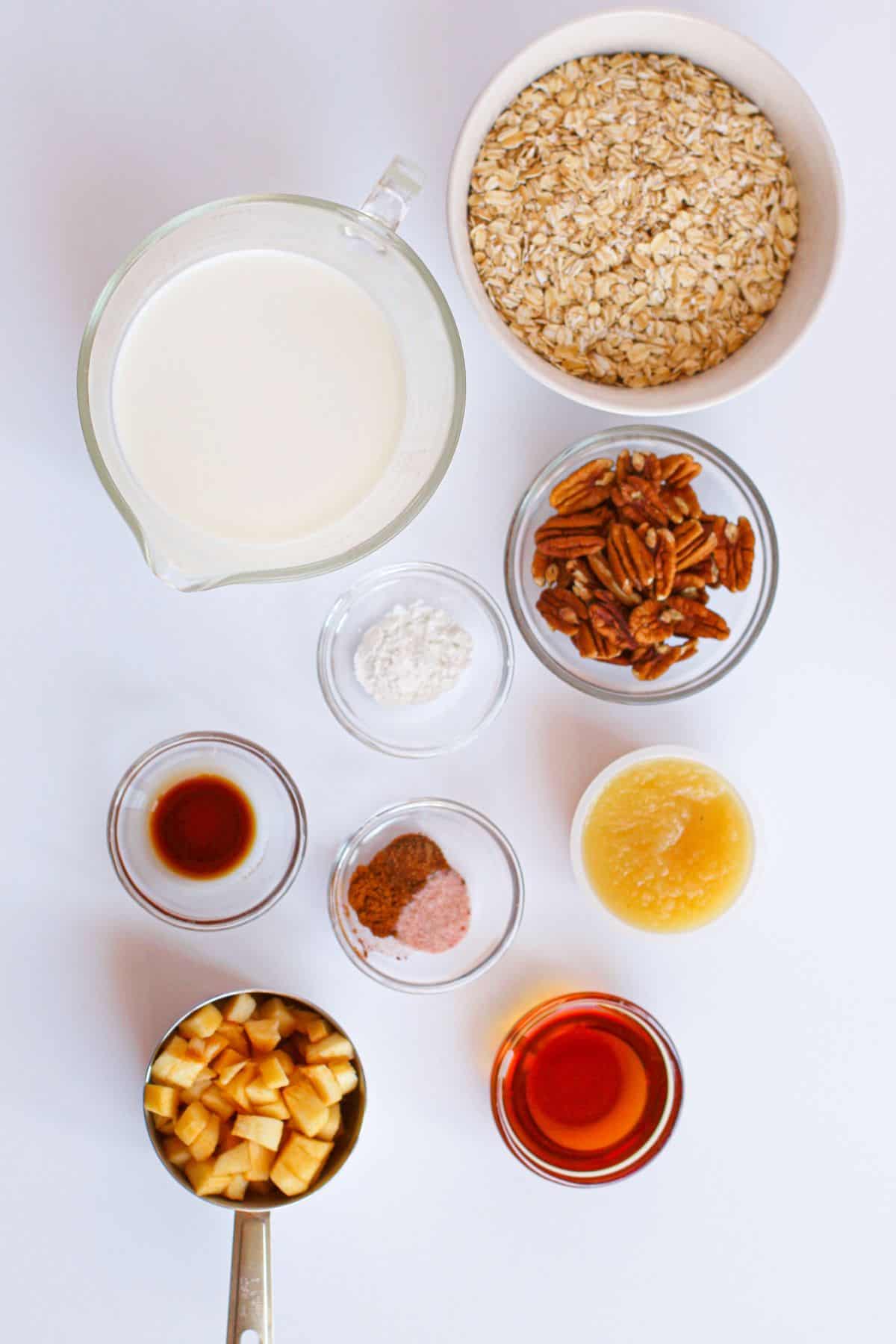 A top-down view of ingredients for Baked Apple Oatmeal in separate bowls and cups, including oats, milk, pecans, apples, applesauce, maple syrup, vanilla, cinnamon, salt, and baking powder—all arranged on a white background.