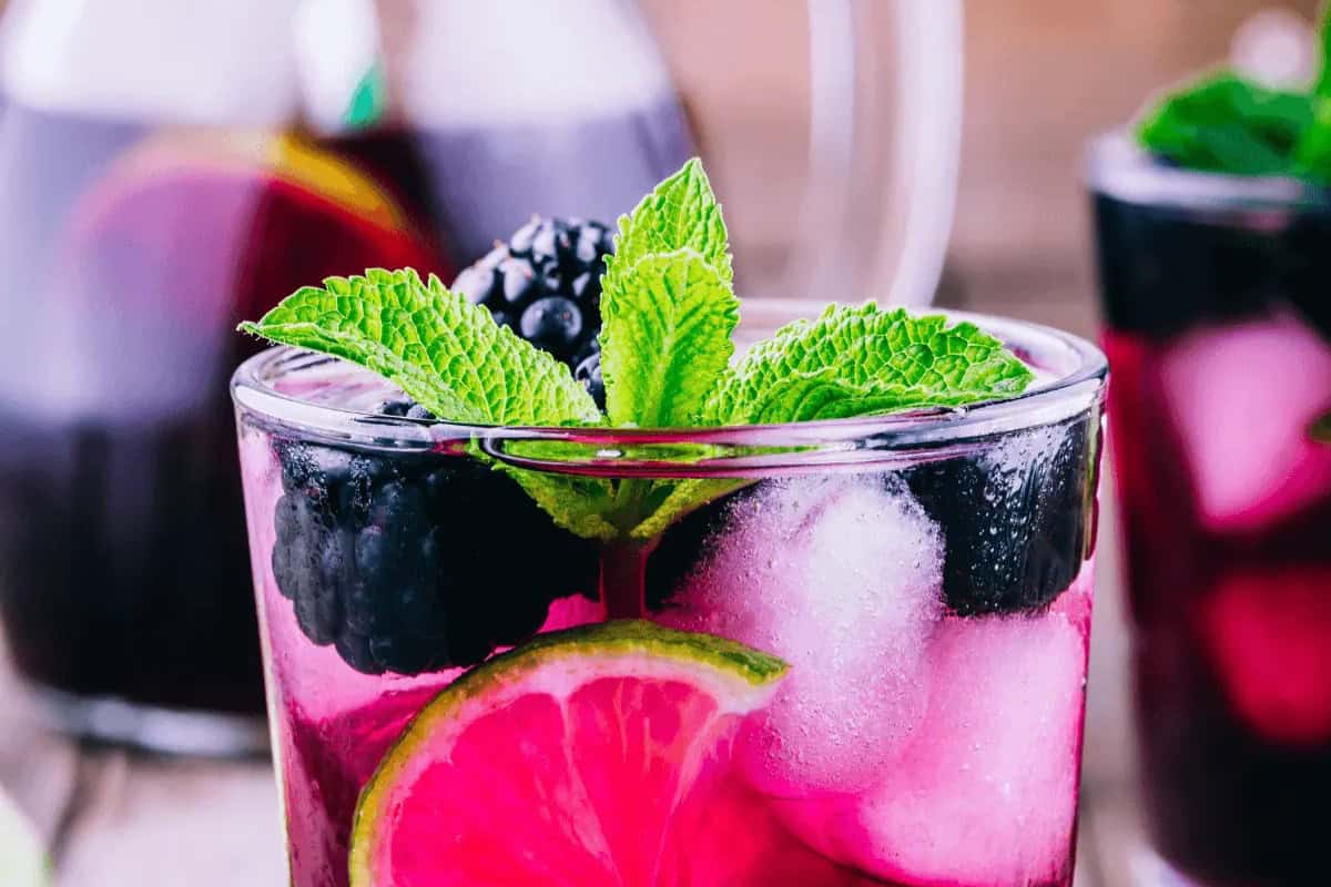 A glass of purple blackberry bourbon smash drink with ice, blackberries, a lime slice, and fresh mint leaves. Additional refreshing drinks are seen in the background.
